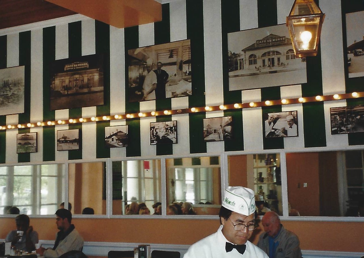 A man in chef 's hat and tie standing at the counter.
