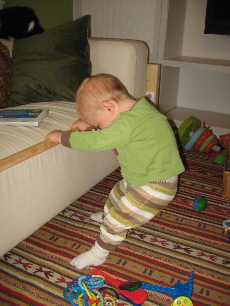 A baby in green shirt and striped pants standing on the floor.