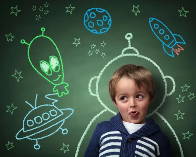 A little boy standing in front of a chalkboard with alien drawings on it.