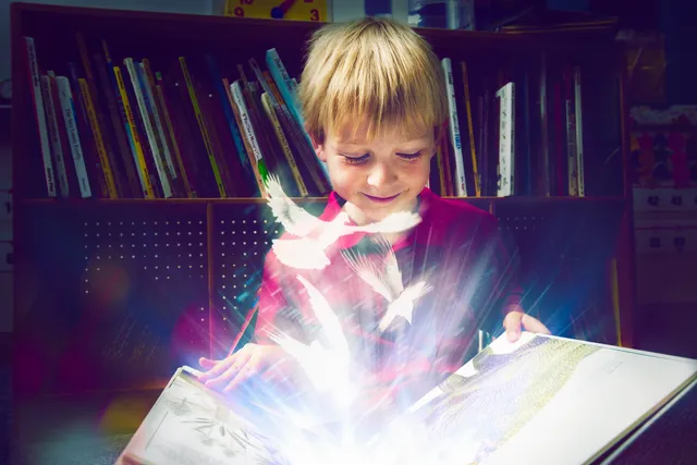 A young boy is looking at the light coming from his book.